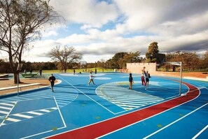 An outdoor basketball court with a blue and red playing surface, surrounded by trees and a cloudy sky. - Sleep 5 | Luxury Box Hill Townhouse (Box Hill)