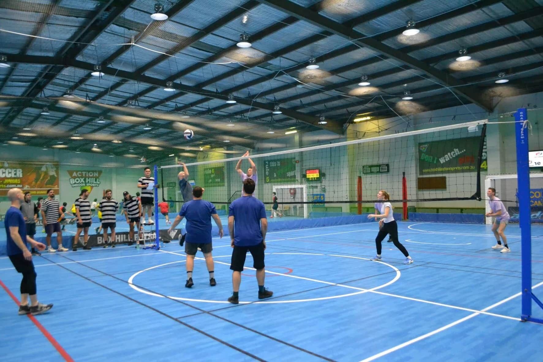 An indoor sports facility with a blue floor, a volleyball net, and spectators.