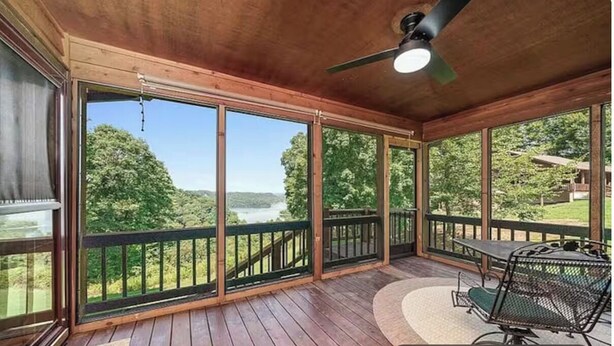 A covered deck with a view of a lake and forest, a ceiling fan, and a table with chairs.