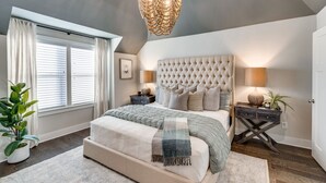 Een slaapkamer met een gestoffeerd hoofdeinde, een groot bed, nachtkastjes, een kroonluchter en een raam met gordijnen.