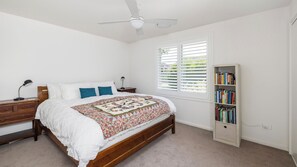 Quarto com cama de madeira, roupa de cama branca, colcha, mesa de cabeceira com abajur, estante de livros e janela com persianas.