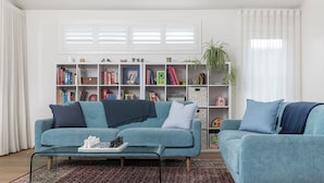 Ein Wohnzimmer mit einem petrolfarbenen Sofa, einem passenden Sessel und einem Couchtisch mit einem Schachspiel. Im Hintergrund steht ein Bücherregal mit Büchern und Dekoration.