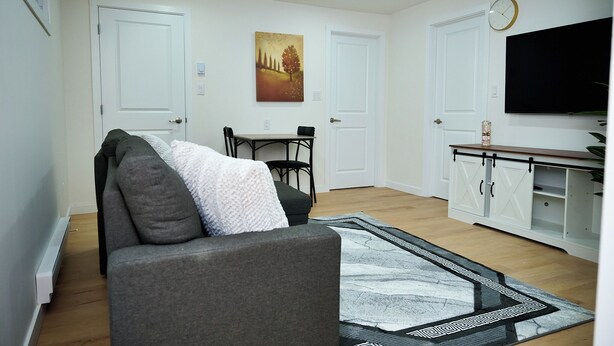A living room with a grey sofa, a dining table with chairs, a painting on the wall, and a TV mounted above a white cabinet.