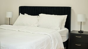 A neatly made bed with white linens and pillows, flanked by two bedside tables with lamps, in a simple room with a plain wall and a dark headboard.