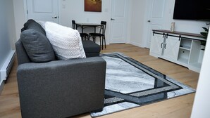 A modern living room with a grey sofa, a dining table with chairs, a flat-screen TV, and a patterned rug.