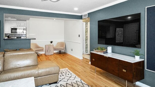 A modern living room with a leather sofa, a wooden floor, a ceiling fan, a TV on the wall, and a kitchen in the background. - Stylish Experience in Buckhead (Atlanta)