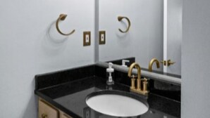 A bathroom with a black countertop, a white sink, gold fixtures, and a mirror with lights above. - Stylish Experience in Buckhead (Atlanta)