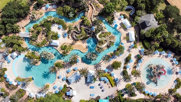 Eine Vogelperspektive zeigt ein Resort mit mehreren Swimmingpools, Liegebereichen und Palmen.