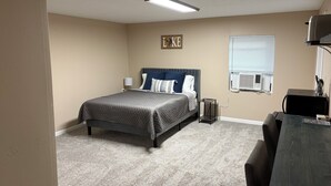 Un dormitorio con cama, mesita de noche, lámpara, aire acondicionado y un escritorio con computadora.