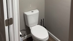 Un baño con inodoro, toallero con toallas dobladas, dispensador de papel higiénico y un pequeño estante con una botella y difusor de aroma.