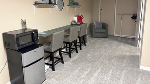 Una cocina compacta con microondas, refrigerador y una pequeña mesa de comedor con sillas. Hay repisas sobre la encimera con platos y un reloj.
