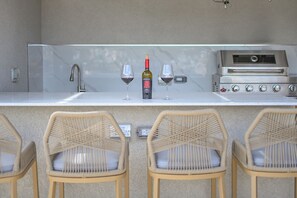 An outdoor bar area with a wine bottle, two glasses, a grill, and wicker chairs. - 10BR Golden Pear Villa-MS, Sea View – Family/Group Ready (Saint George)