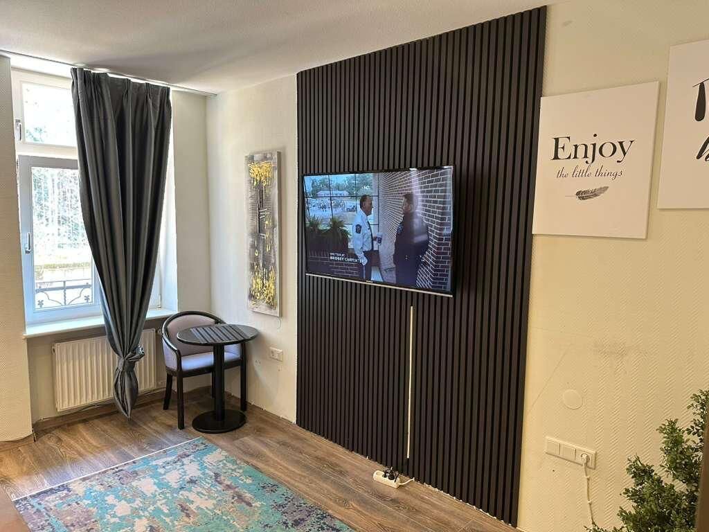 A room with a television mounted on a wall, a window with curtains, a chair, and a rug.