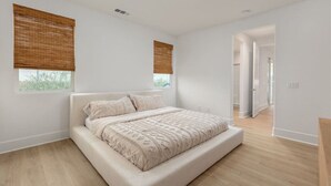 Ein Schlafzimmer mit einem großen Bett, zwei Fenstern mit Jalousien, Holzboden und einem zur anderen Kammer führenden Flur.