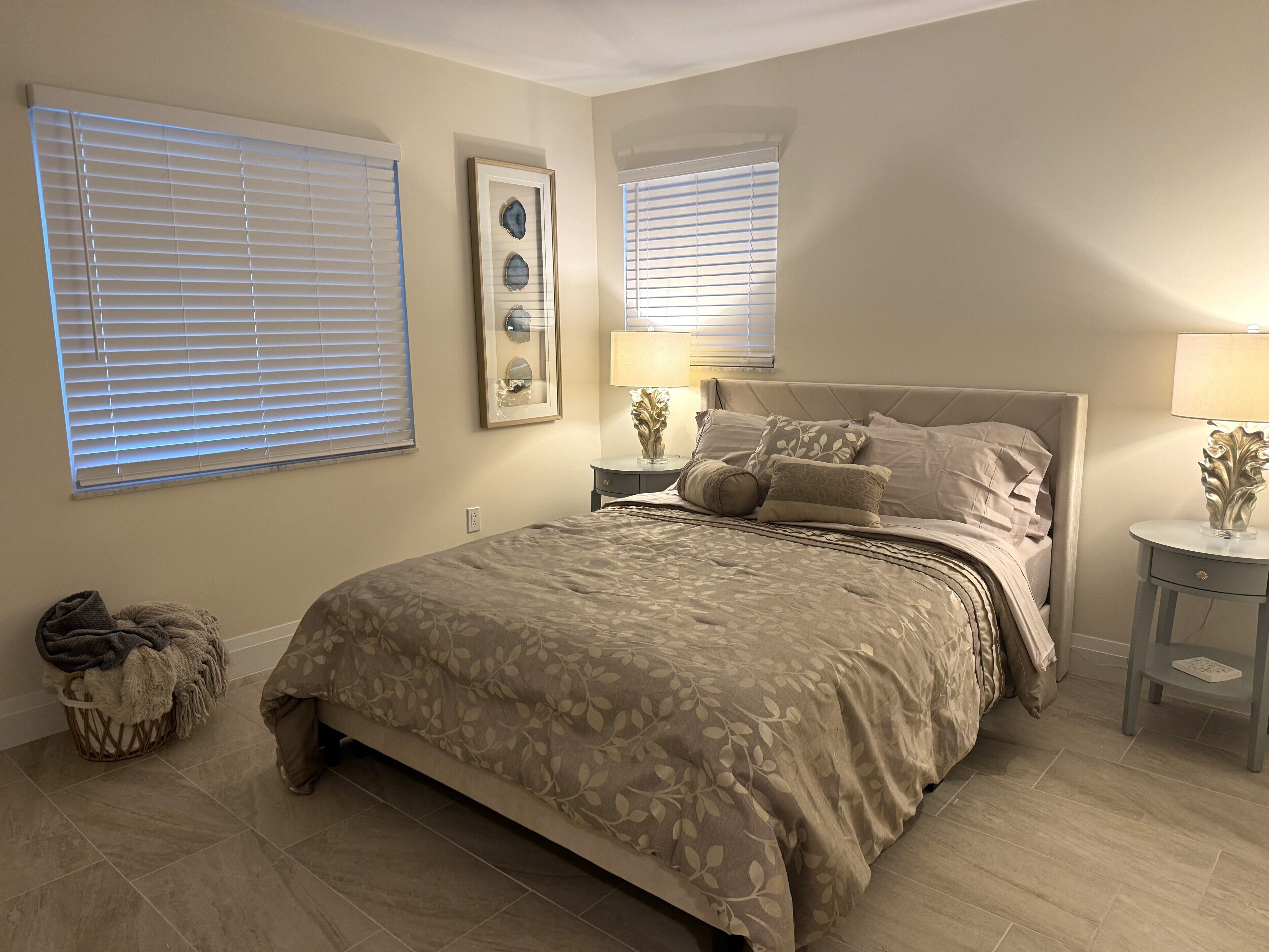A bedroom with a bed, two nightstands, a lamp, a framed picture, and a window with blinds.