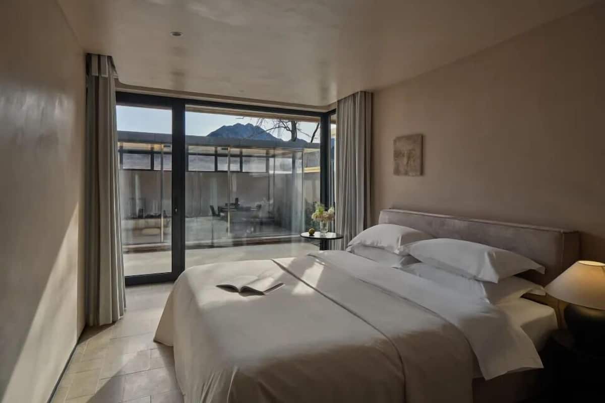 A hotel room with a large bed, a bedside table with a lamp, and a view of a mountain through a glass door.