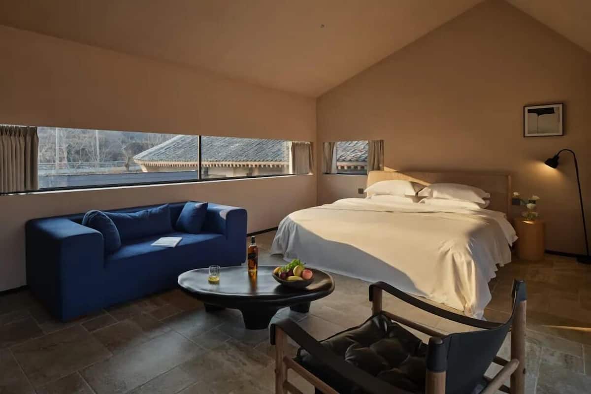 A bedroom with a bed, a blue sofa, a small table with a bottle and fruit, and a chair with a cushion.
