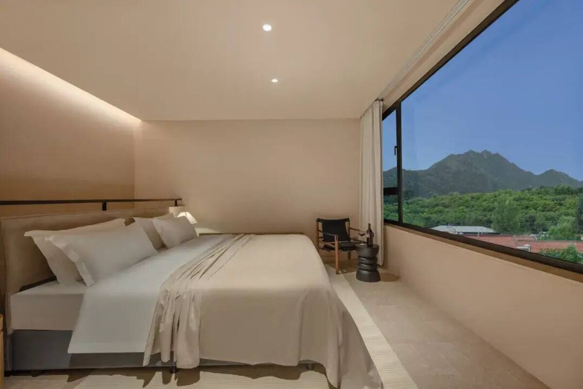 A modern bedroom with a large bed, a small table, and a view of mountains through a large window.