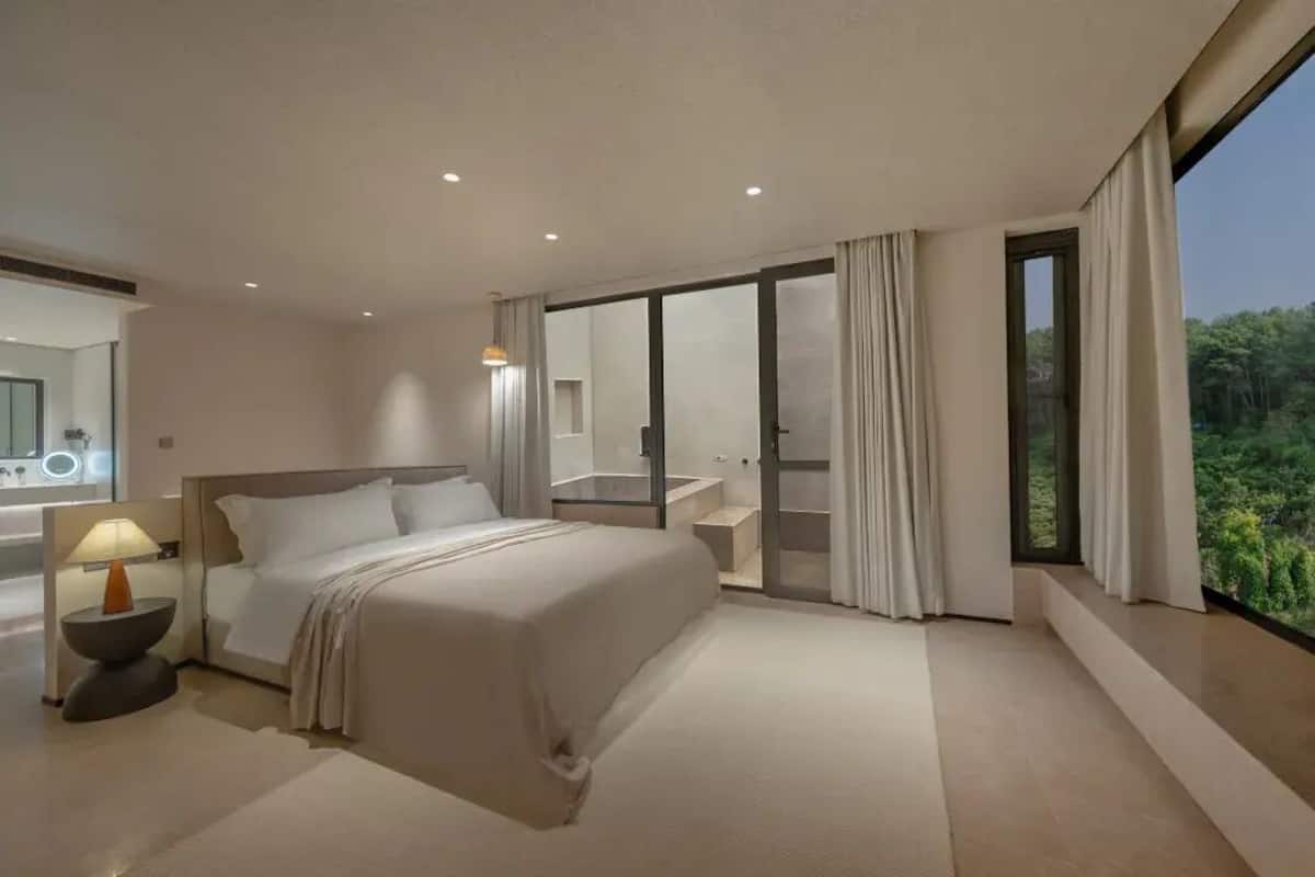 A modern hotel bedroom with a large bed, a bedside table with a lamp, a bathroom with a bathtub, and a sliding glass door leading to a balcony with a view of greenery.