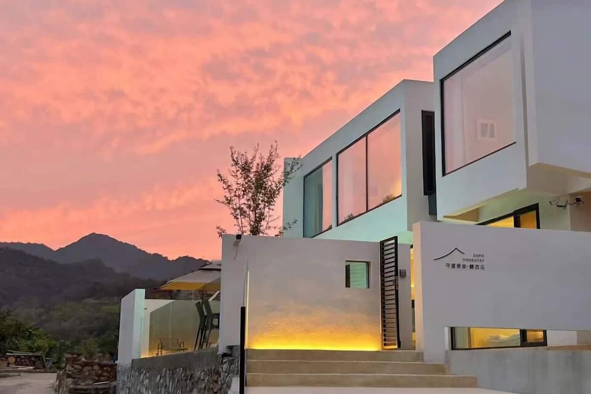A modern two-story building with large windows and a flat roof, set against a mountainous backdrop during sunset.