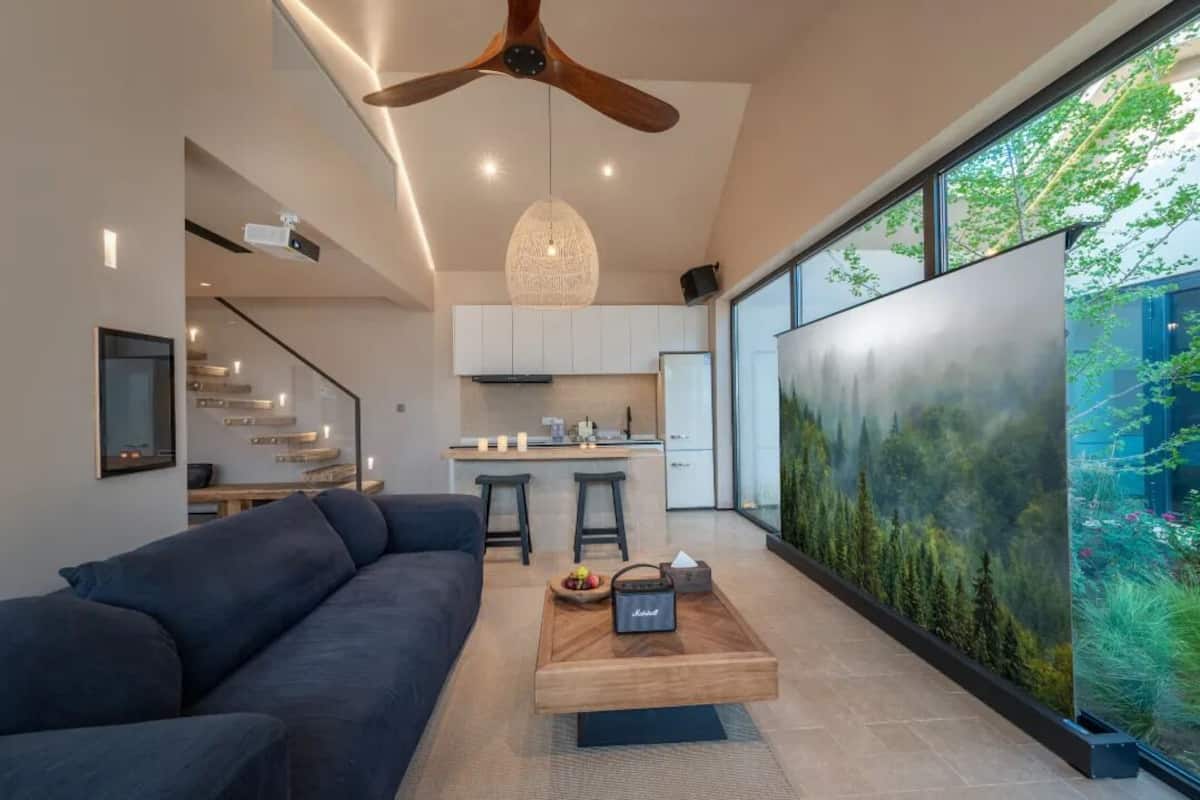 A modern living room with a dark sofa, a wooden coffee table, and a large window with a forest mural.