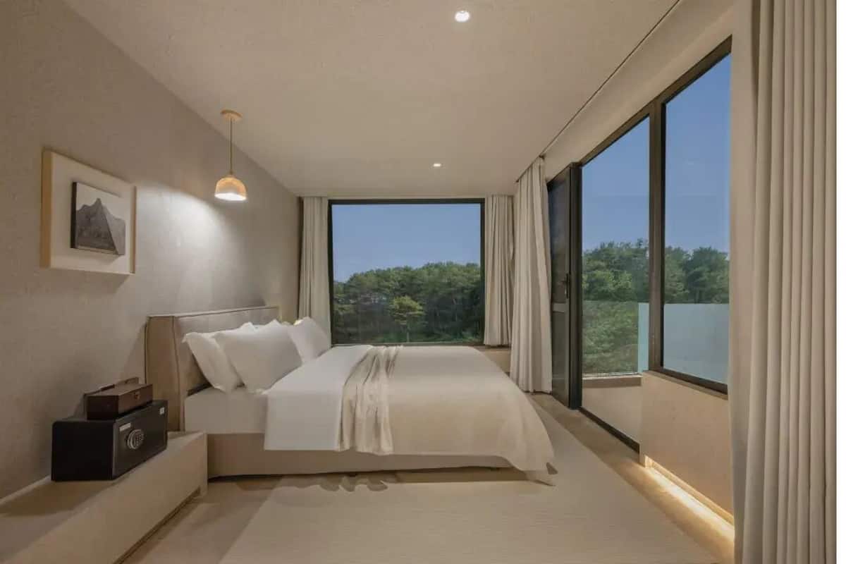 A modern bedroom with a large bed, a nightstand, and a view of greenery through large windows.