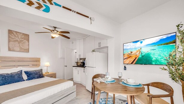 Habitació d'hotel moderna amb un llit, zona de dinar amb taula i cadires, ventilador de sostre, cuina i televisió que mostra una escena de platja.