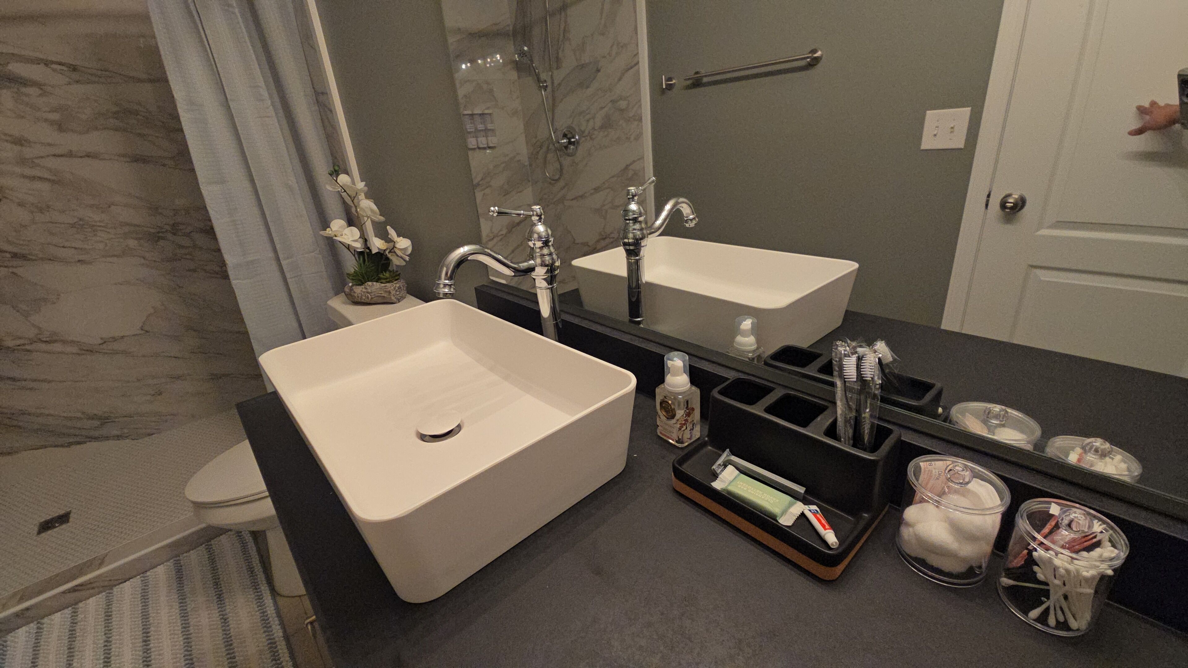 A bathroom with a large white sink, a mirror, and a shower. There are toiletries and a towel rack visible.