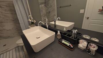 A bathroom with a large white sink, a mirror, and a shower. There are toiletries and a towel rack visible.
