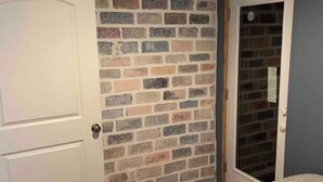 A room with a brick-patterned wall, a white door, a mirror, and a bed with a grey blanket. - 8 miles From IU Campus: Country & Quiet  2-BR apt  in charming Ellettsville (Ellettsville)