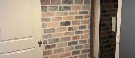 A room with a brick-patterned wall, a white door, a mirror, and a bed with a grey blanket.