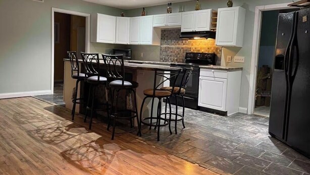 A kitchen with a central island, bar stools, white cabinets, a black refrigerator, and a tiled backsplash. - 8 miles From IU Campus: Country & Quiet  2-BR apt  in charming Ellettsville (Ellettsville)