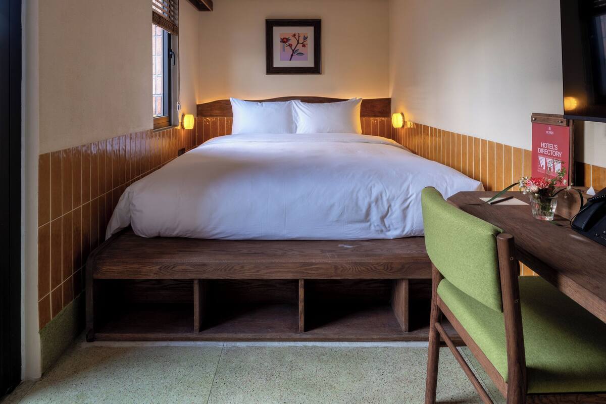 A hotel room with a wooden bed frame, a green chair, a desk with a telephone, and a framed picture on the wall.