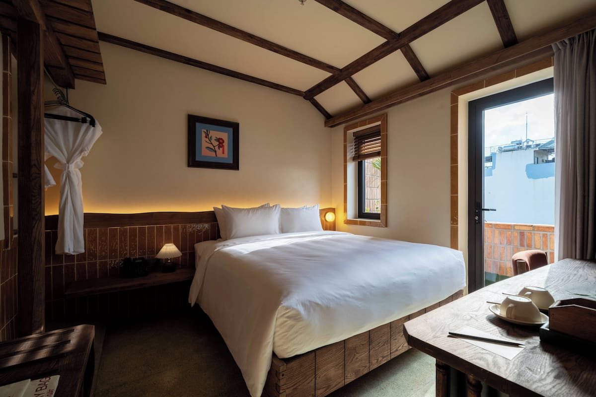 A bedroom with a wooden bed, a bedside table, a hanging robe, a framed picture, and a window with blinds.