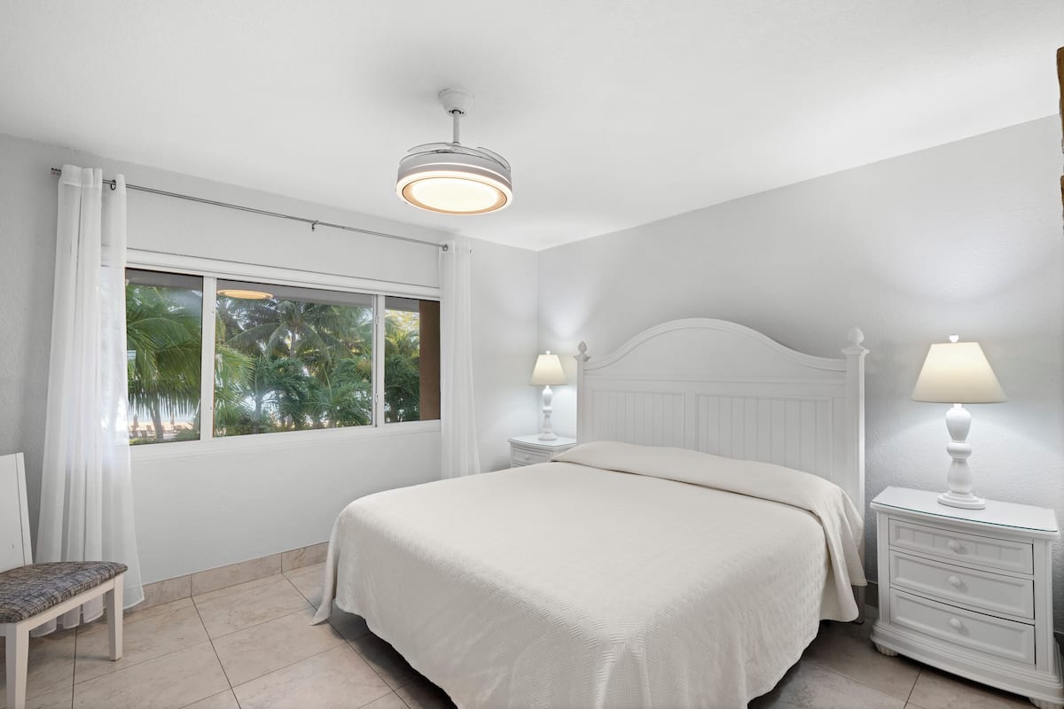 Un dormitorio con una cama grande, dos mesitas de noche con lámparas, una silla y una ventana con cortinas.