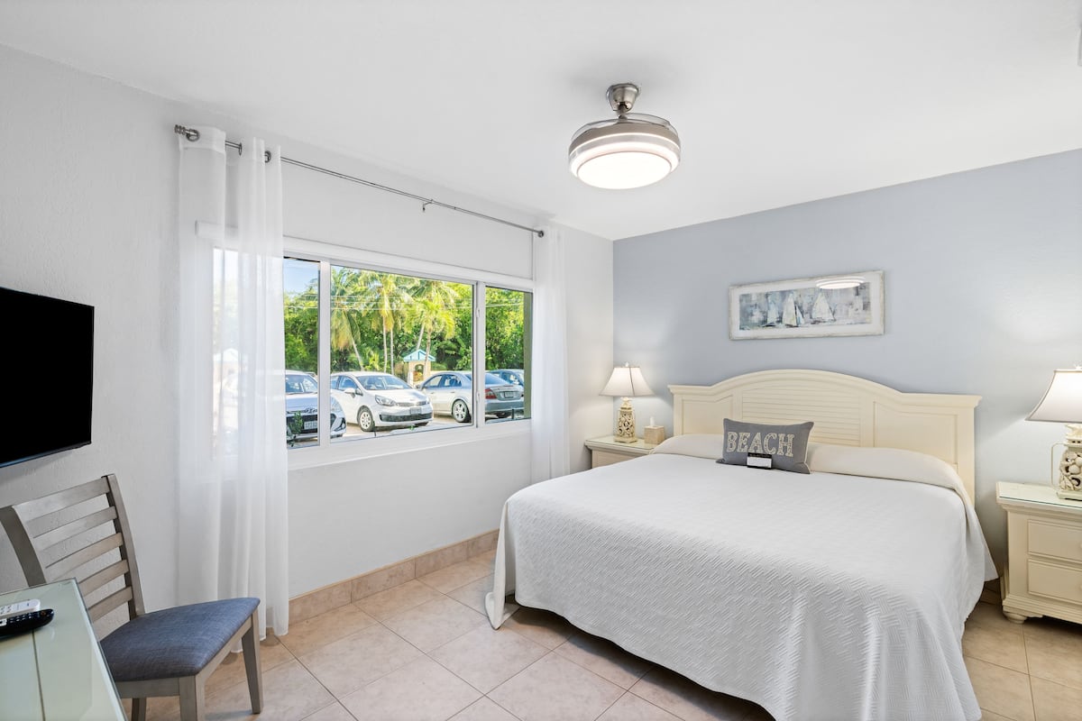 Habitación de hotel con cama, televisor, sillón, ventana con vistas a coches aparcados y palmeras, y ventilador de techo.