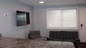 Ein Zimmer mit Bett, Wandfernseher, Fenster mit Jalousien, Sofa und Tür.