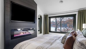 Une chambre moderne avec une cheminée, une grande fenêtre donnant sur la forêt et un lit agrémenté d’une literie à motifs.
