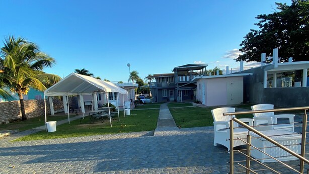 A paved pathway leading to a covered area with white chairs, a palm tree, and a modern building with a garage. - Cozy 1-bedroom apartment in beautiful Aguada with AC (Aguada)