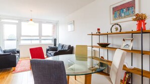 A modern living room with a glass-top coffee table, a red chair, and a black sofa. There is a bookshelf with decorative items and a framed picture on the wall. - Bright & Spacious 2 Bed Islington Flat | Fast Wi-Fi (London)