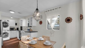 A modern kitchen with white cabinetry, a washing machine, and a dining area with a wooden table and chairs. - Bungalow + Casita With Private Patios Near Downtown / U of A (Tucson)