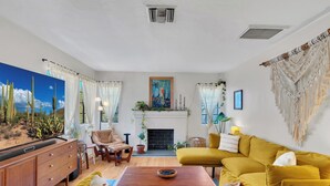 A living room with a sofa, a wooden coffee table, a fireplace, and a cactus painting. - Bungalow + Casita With Private Patios Near Downtown / U of A (Tucson)