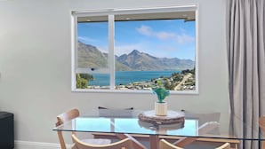 Un comedor con una mesa de vidrio y sillas de madera, una ventana que ofrece vistas a montañas y un lago, y una cortina gris a la derecha.