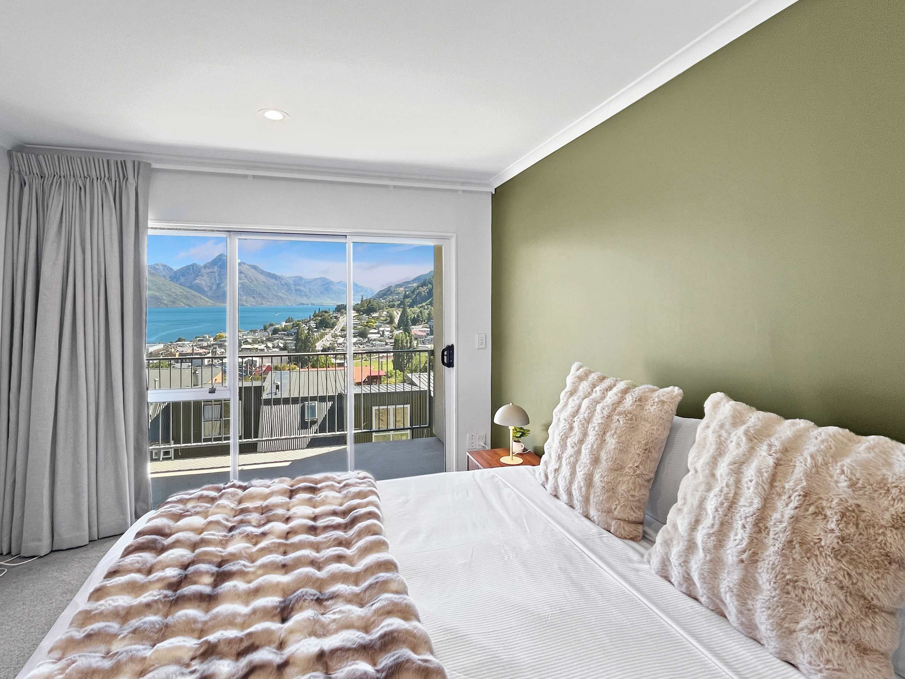 A bedroom with a large bed, a view of a lake and mountains, and a balcony with a railing.