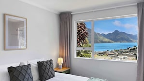 Una habitación de hotel con una cama, una mesita de noche con una lámpara, un cuadro en la pared y una ventana con vistas a montañas y un pueblo.