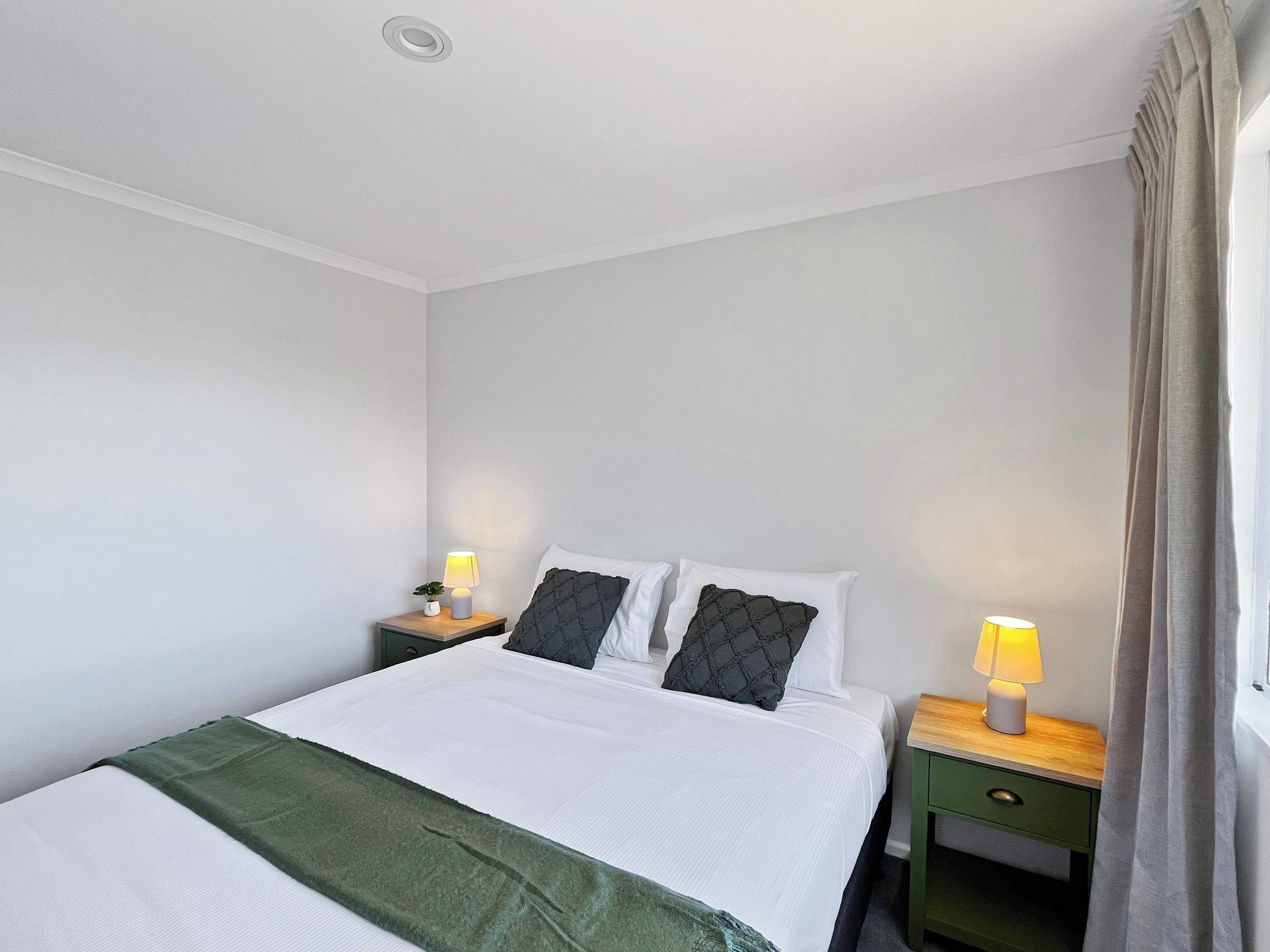 Ein ordentlich gemachtes Bett mit weißen Bettwäsche und einer grünen Tagesdecke, flankiert von zwei Nachttischen mit Lampen, in einem Zimmer mit schlichten weißen Wänden und einem Fenster mit Vorhängen.