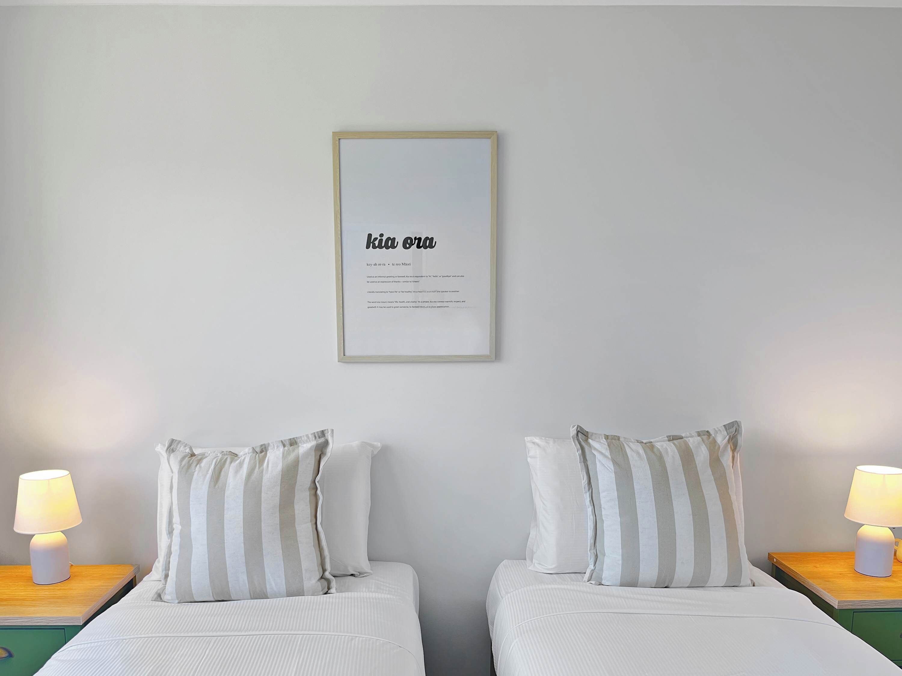 A hotel room with two beds, each with striped pillows and a bedside lamp. A framed quote hangs on the wall above the beds.