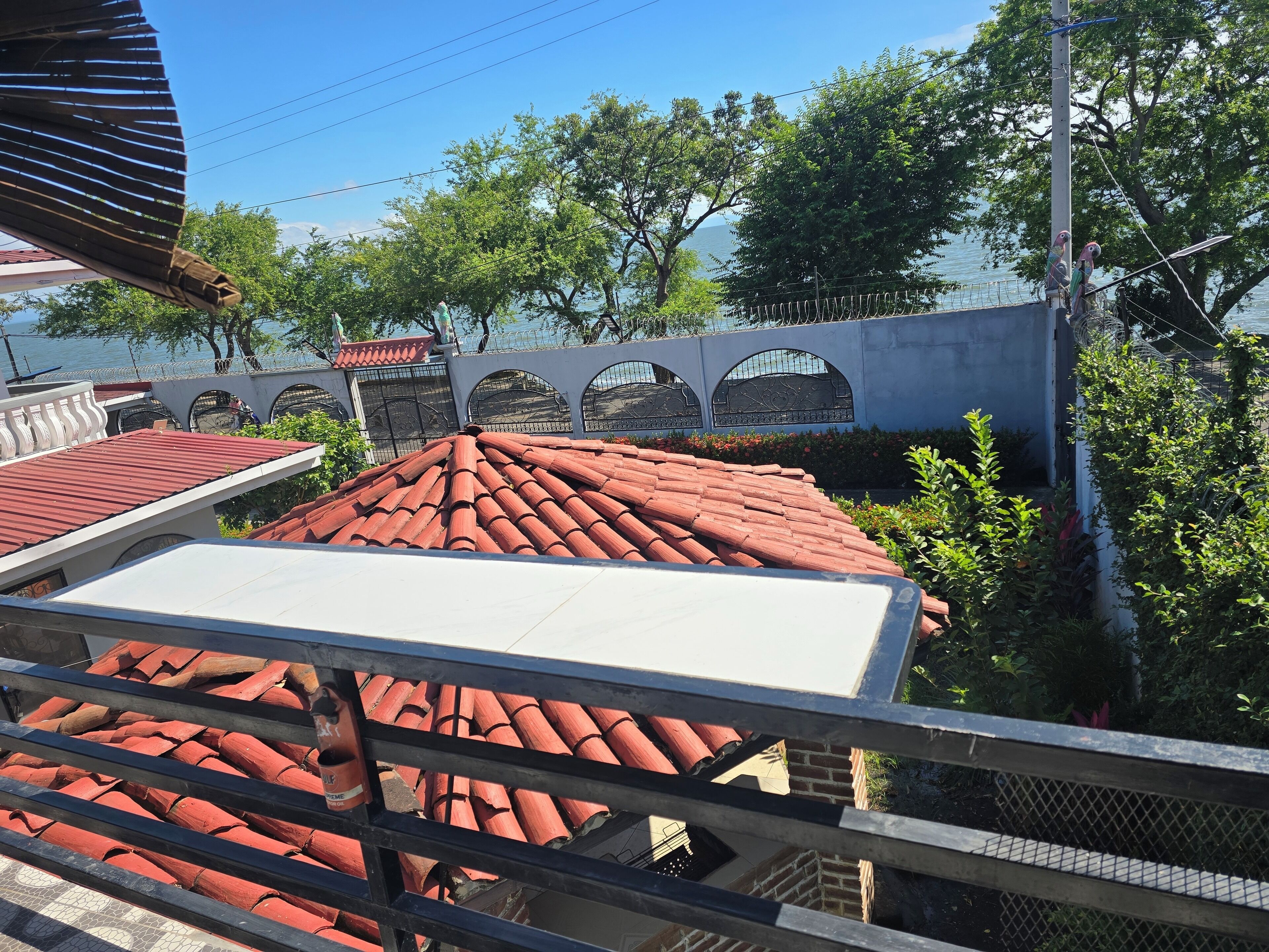 Vista de um edifício com telhado de telhas vermelhas, varanda branca e grade de metal. Árvores e uma cerca aparecem ao fundo.