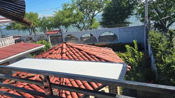 Vista de um edifício com telhado de telhas vermelhas, varanda branca e grade de metal. Árvores e uma cerca aparecem ao fundo.