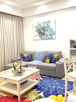 A living room with a grey sofa, colorful pillows, a glass coffee table, and a floral carpet. - Juan Dolio Executive One Bedroom Suite With Ocean Views. (Juan Dolio)
